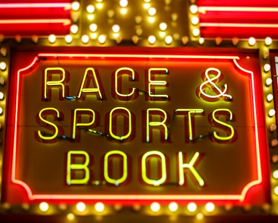 Bright neon lights on a marquee sign display the words 'RACE & SPORTS BOOK'. The sign is surrounded by rows of yellow and white lights against a red background, evoking a vibrant, energetic atmosphere typically associated with casinos or entertainment venues.