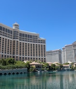 A large luxury hotel and casino building with multiple stories, featuring a light facade with arched windows. The structure has a prominent central tower with a dome-like top. It is surrounded by landscaping with trees and a body of water in the foreground.