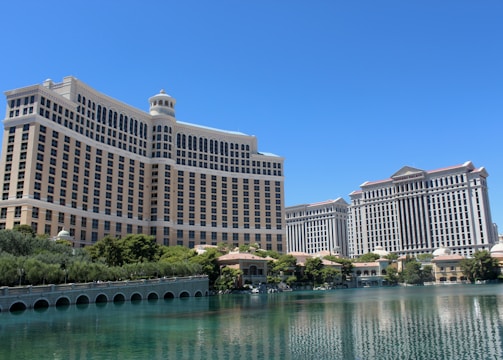 A large luxury hotel and casino building with multiple stories, featuring a light facade with arched windows. The structure has a prominent central tower with a dome-like top. It is surrounded by landscaping with trees and a body of water in the foreground.