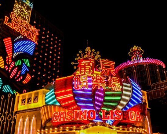 Bright neon lights and colorful signage adorn the exterior of Casino Lisboa, creating a vibrant and lively atmosphere. The façade features bold, multicolored designs with a mix of English and Chinese characters prominently displayed. The lights form intricate patterns and shapes, contributing to the energetic ambiance of the scene.