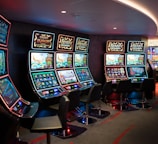 A row of brightly lit slot machines in a casino setting, each with colorful displays and various game themes. The machines are lined up against a dark wall and are unoccupied, with cushioned chairs placed in front of them. The area is dimly lit except for the glow from the screens, creating a vibrant and engaging atmosphere.