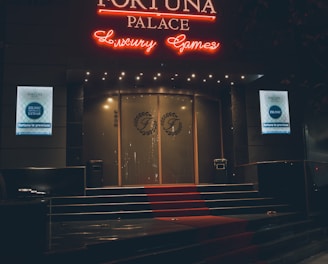 A neon-lit casino entrance with a red carpet leading up to large double doors. The sign reads 'Fortuna Palace Luxury Games' in bright red and white neon lights. The dimly lit atmosphere suggests a night-time setting. There are two digital displays on either side of the entrance showing promotional content.
