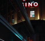 A night scene featuring a building with illuminated neon signage displaying 'CASINO'. The environment is urban with reflections on glass windows and glowing street traffic lights below. The atmosphere is dimly lit, with artificial lighting casting vibrant colors.