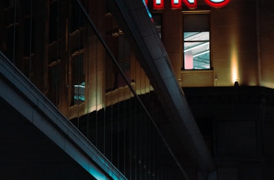 A night scene featuring a building with illuminated neon signage displaying 'CASINO'. The environment is urban with reflections on glass windows and glowing street traffic lights below. The atmosphere is dimly lit, with artificial lighting casting vibrant colors.
