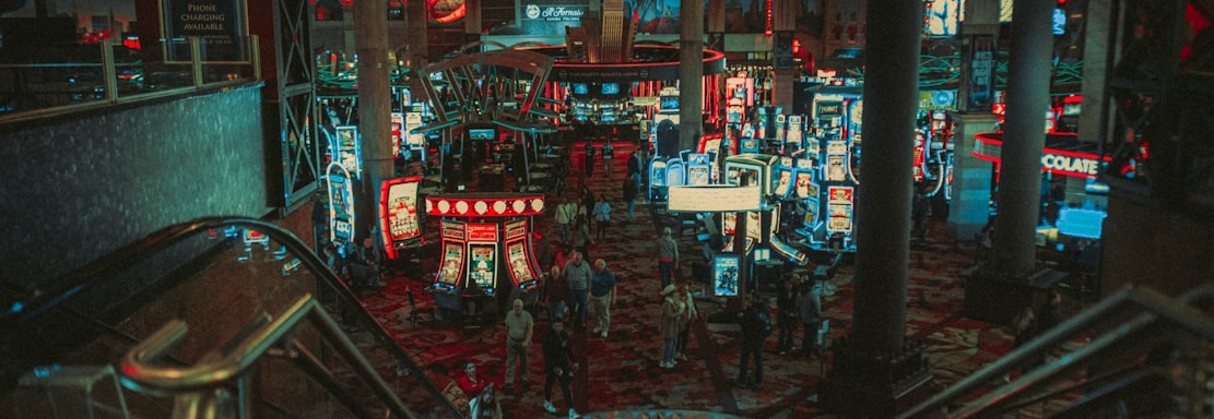 A bustling casino floor filled with groups of slot machines and people walking around. The atmosphere is vibrant with neon lights and illuminated signs. The interior has a ceiling supported by large pillars and there are stairs leading down to the gaming area.