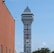A tall tower with a modern design features the large vertical red letters spelling 'CASINO'. The structure has a sleek, metallic appearance with various horizontal and vertical lines. The sky is mostly clear with a few clouds. In the foreground, part of a brick building is visible, along with some trees and buildings in the distance.