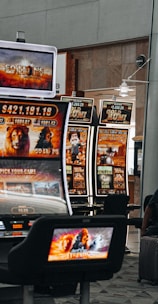 A collection of slot machines is situated inside a building with bright, colorful screens displaying various games and gaming themes. A person is seated nearby, seemingly relaxed with luggage beside them, and the atmosphere is lively with electronic displays.