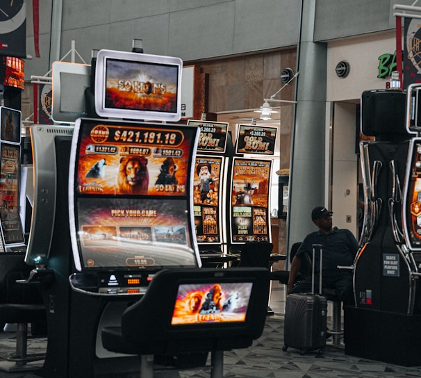 A collection of slot machines is situated inside a building with bright, colorful screens displaying various games and gaming themes. A person is seated nearby, seemingly relaxed with luggage beside them, and the atmosphere is lively with electronic displays.