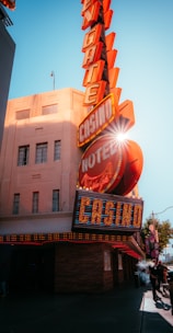 A vibrant city scene featuring a tall, illuminated casino sign with retro-style neon lights. The sign dominates the building facade with the words 'CASINO' and 'HOTEL'. Sunlight peeks through, creating a dramatic lens flare effect. The street is lively, with pedestrians casually walking and a hint of urban hustle.