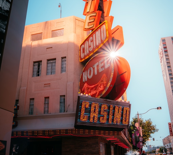 A vibrant city scene featuring a tall, illuminated casino sign with retro-style neon lights. The sign dominates the building facade with the words 'CASINO' and 'HOTEL'. Sunlight peeks through, creating a dramatic lens flare effect. The street is lively, with pedestrians casually walking and a hint of urban hustle.